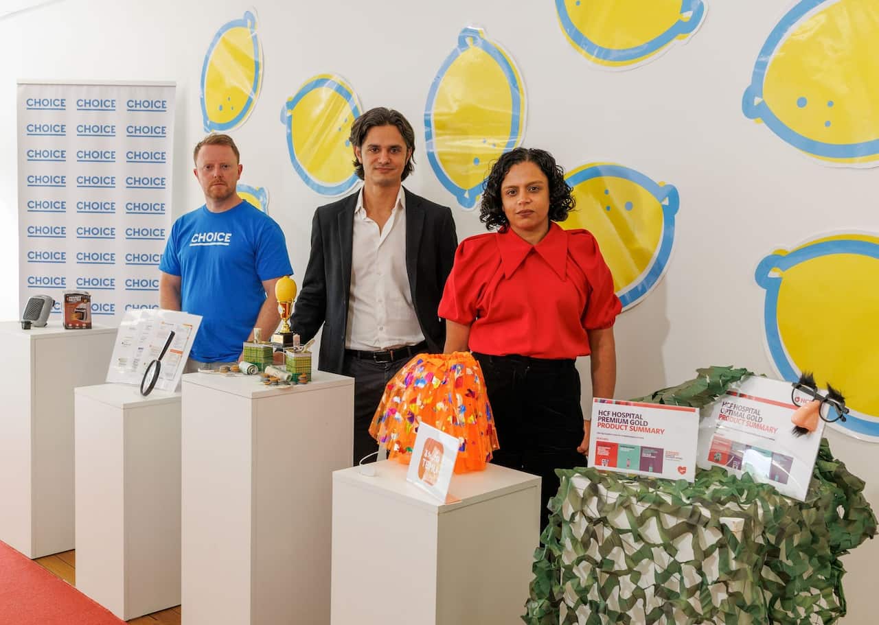 A man in a blue shirt, a man in a white shirt and black blazer, and a woman in a red top. They are standing in front of several white column that have displays.