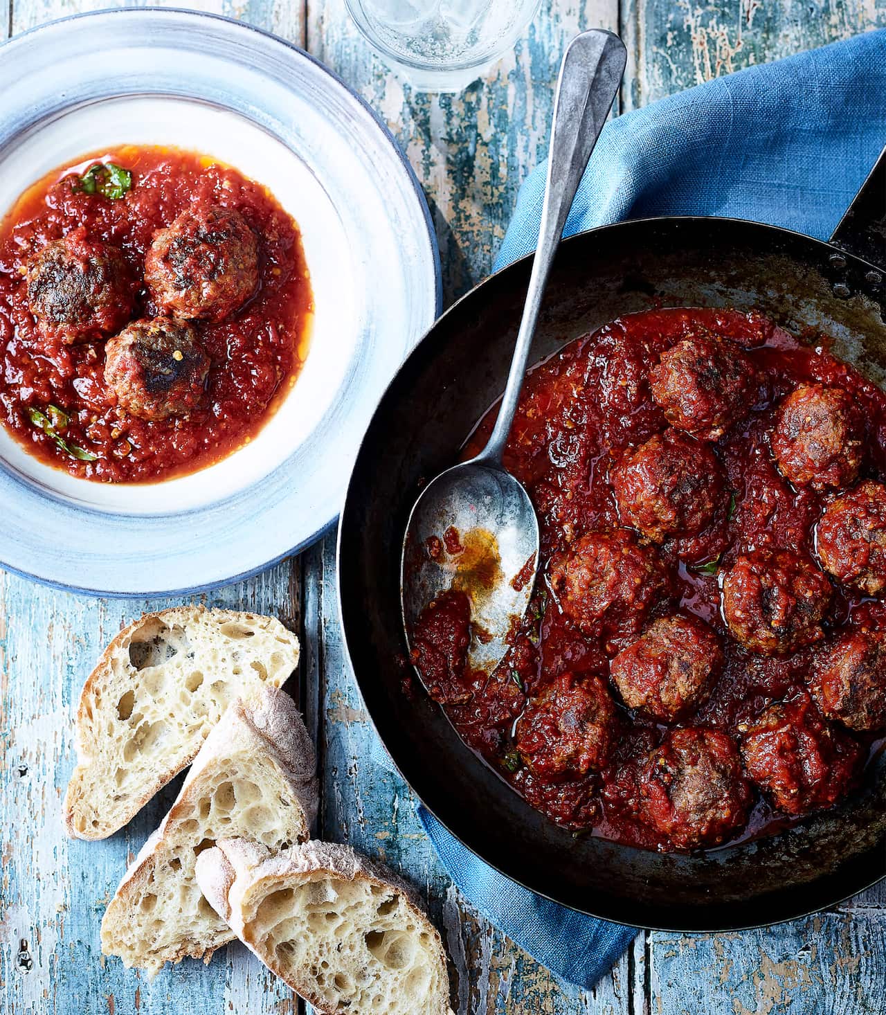A cast iron saucepan sits on a blue napkin, on a blue-painted wooden surface. The pan is full of meatballs in a rich tomato sauce. A pale blue plate sits beside the pan, with a serve of meatballs. Three slices of fresh bread sit nearby.