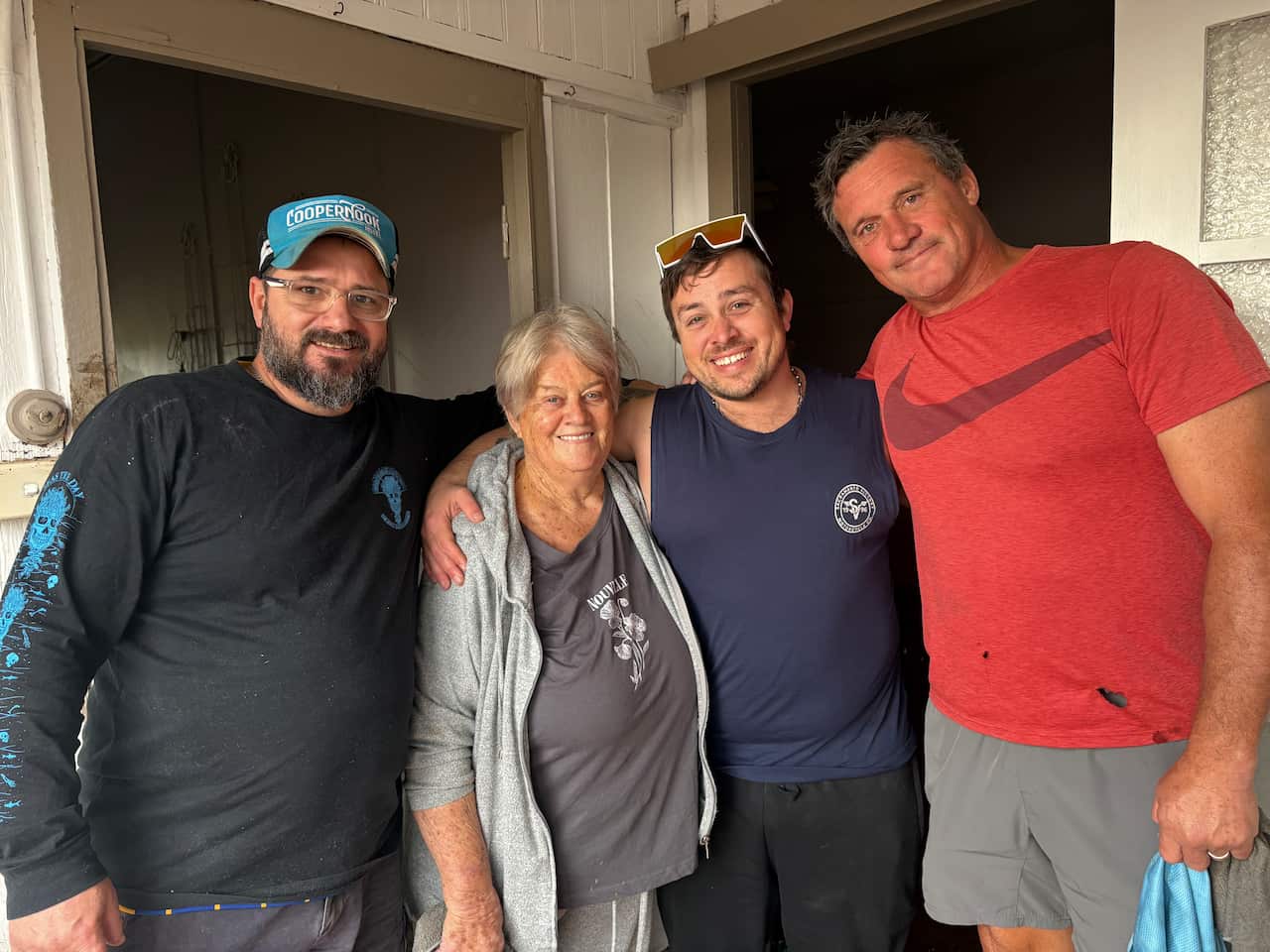 Four people smiling and posing for a photo, standing close together, a middle aged man in a dark long sleeved shirt, an older woman in grey jumper, shirt and track pants, a younger man in a blue singlet, and another tall middle aged man in a coral t-shirt.