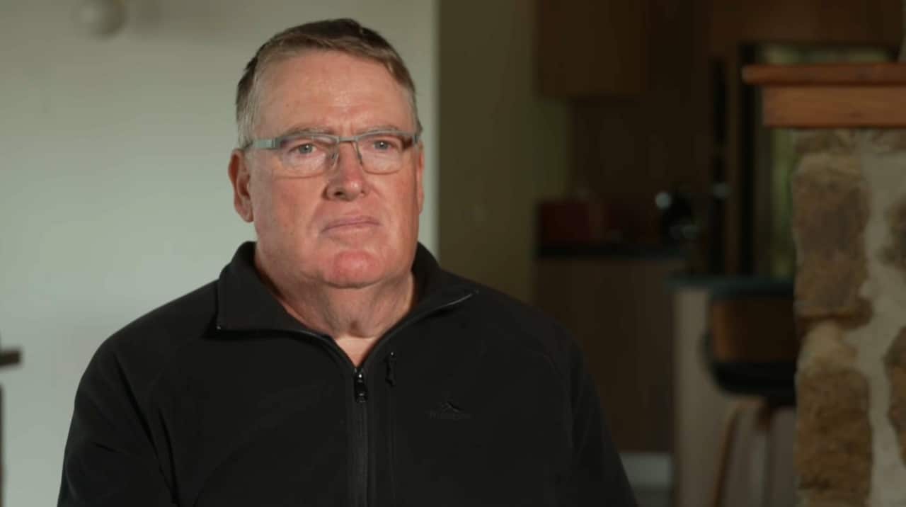 A man wearing a black zip-up top is sitting in a living room.