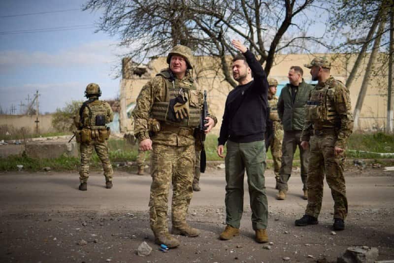 Ukrainian President Volodymyr Zelenskyy standing with troops in Avdiivka, while an armed soldier keeps watch behind them.