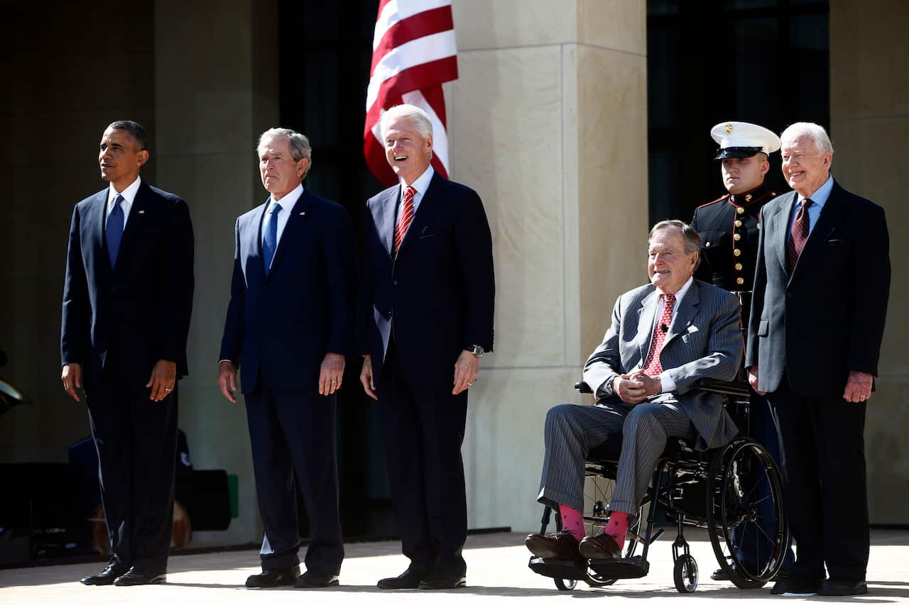 Five men wearing suits and ties pose for a photo, with one man sitting in a wheelchair.