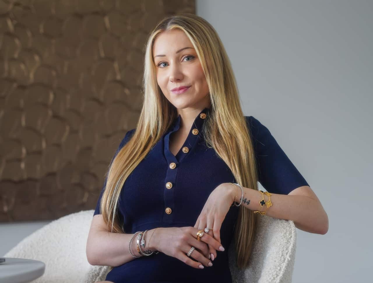 A blonde woman in a blue dress sitting in a chair looking at the camera.