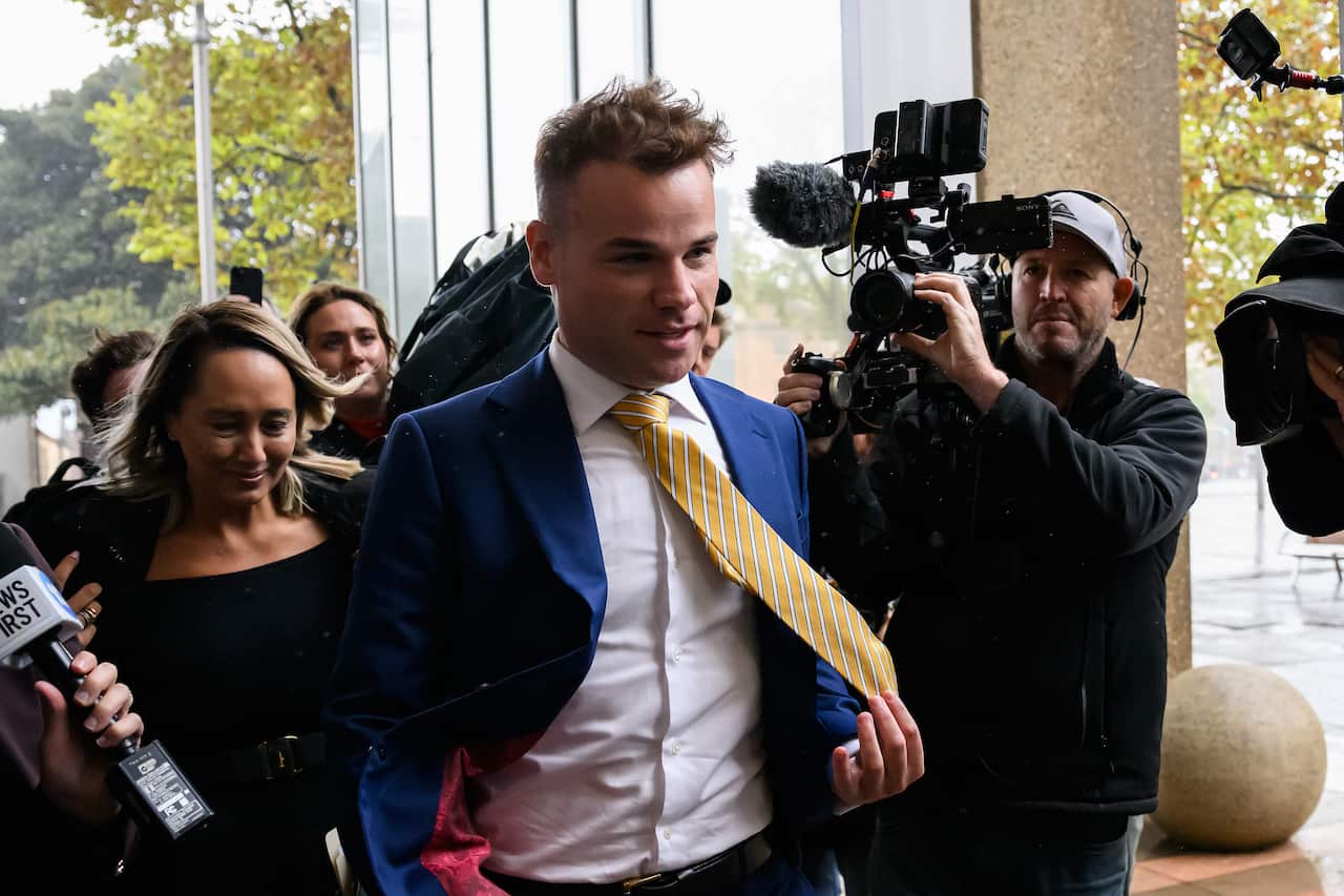 Taylor Auerbach in blue suit and yellow tie leaving court surrounded by media