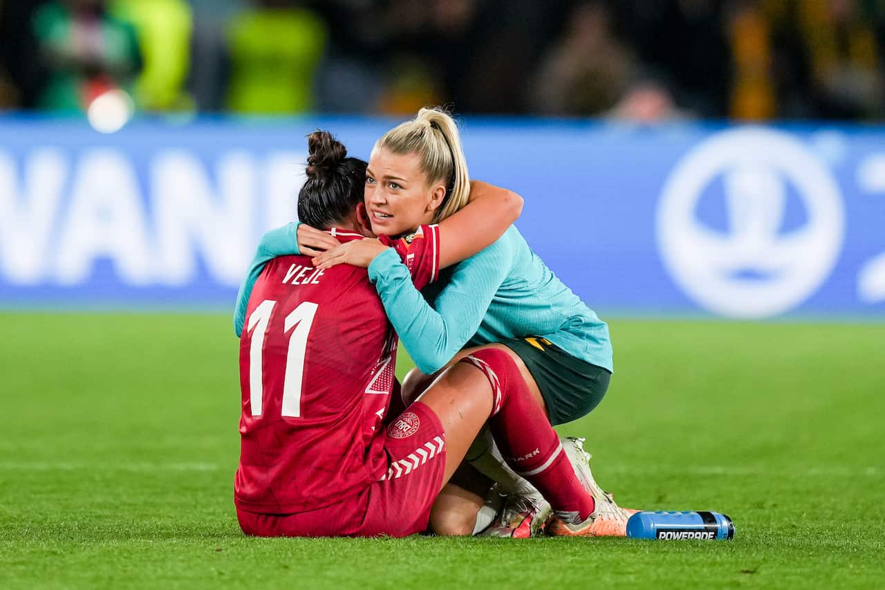 A woman in a teal jacket hugs a woman sitting on the ground in a red football uniform