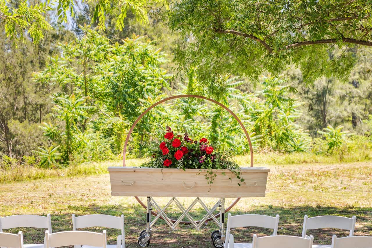 A coffin with red flowers on top stands outside in a park.