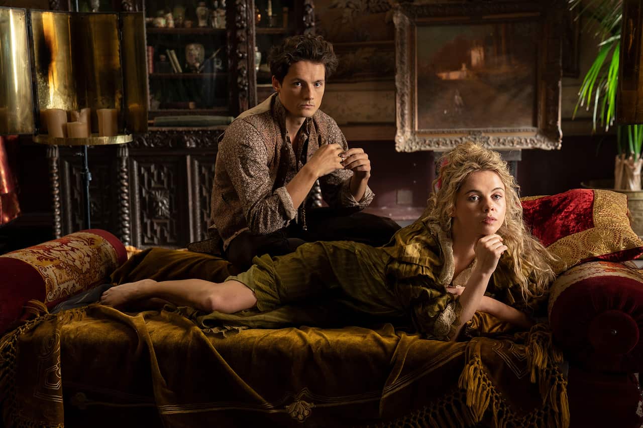 In a sumptuous 19th century setting, a  woman with flowing blonde hair, clothed in a loose wrap, lies on a couch. A yound man sits behind the couch. 