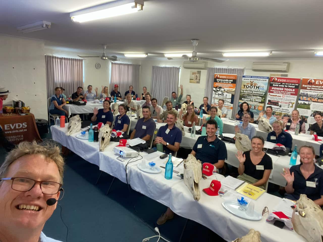 A man in glasses smiling and taking a self-portrait in front of a class of his students