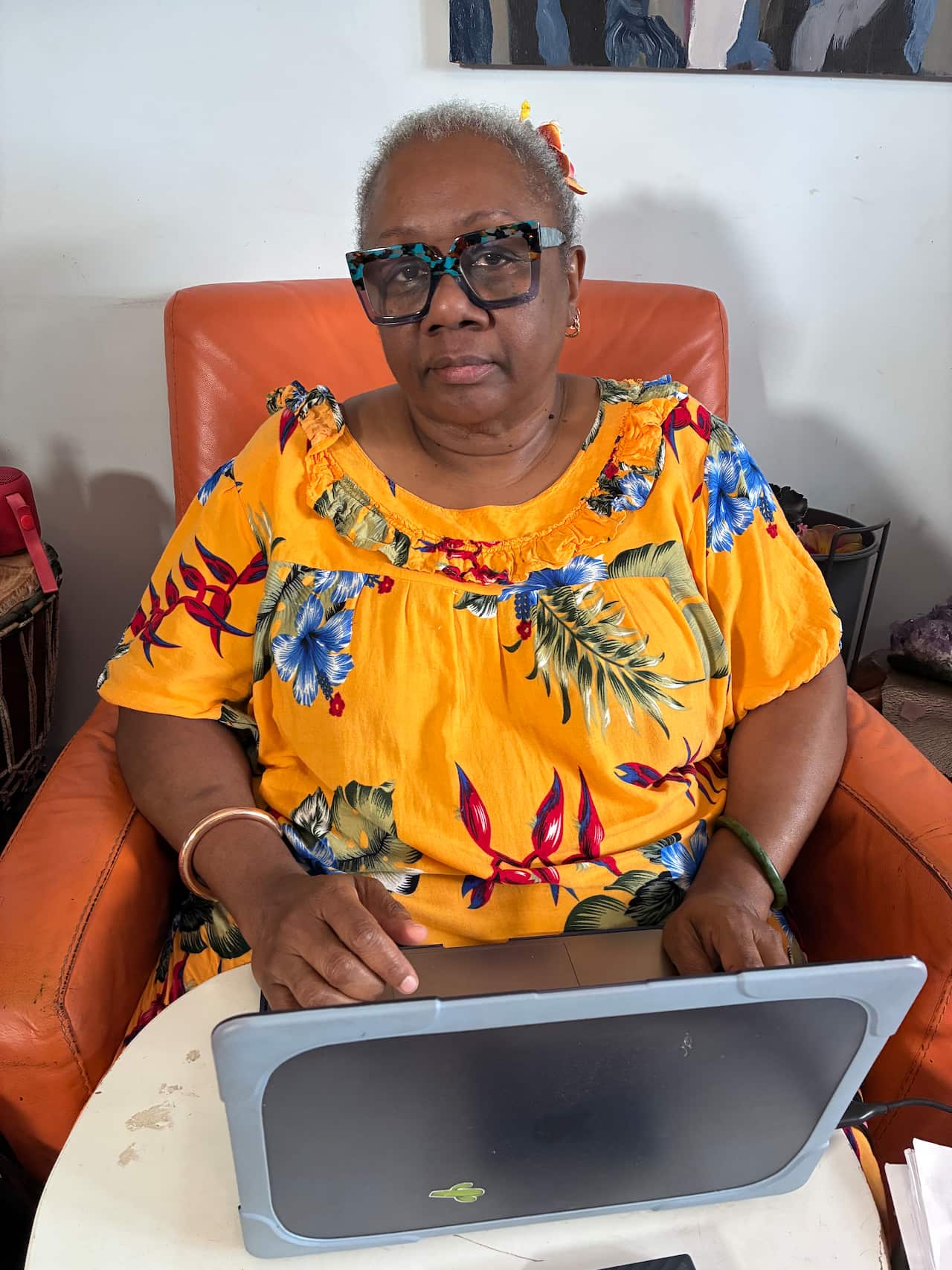 A South Sea Islander woman wearing a colourful yellow dress sits in a large orange chair in front of a laptop.