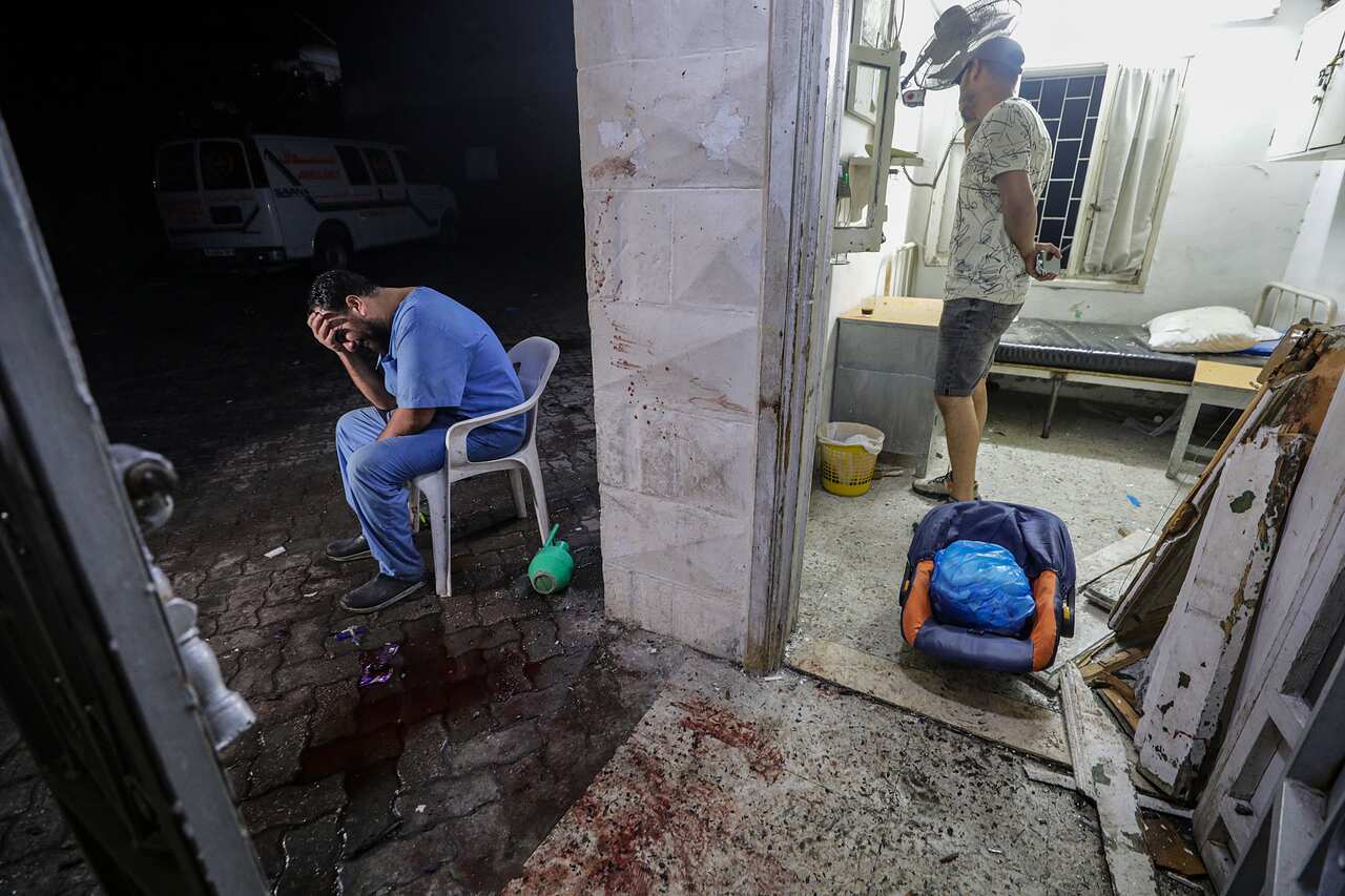 A man in blue scrubs sits on a chair outside a damaged hospital with his hand over his eyes