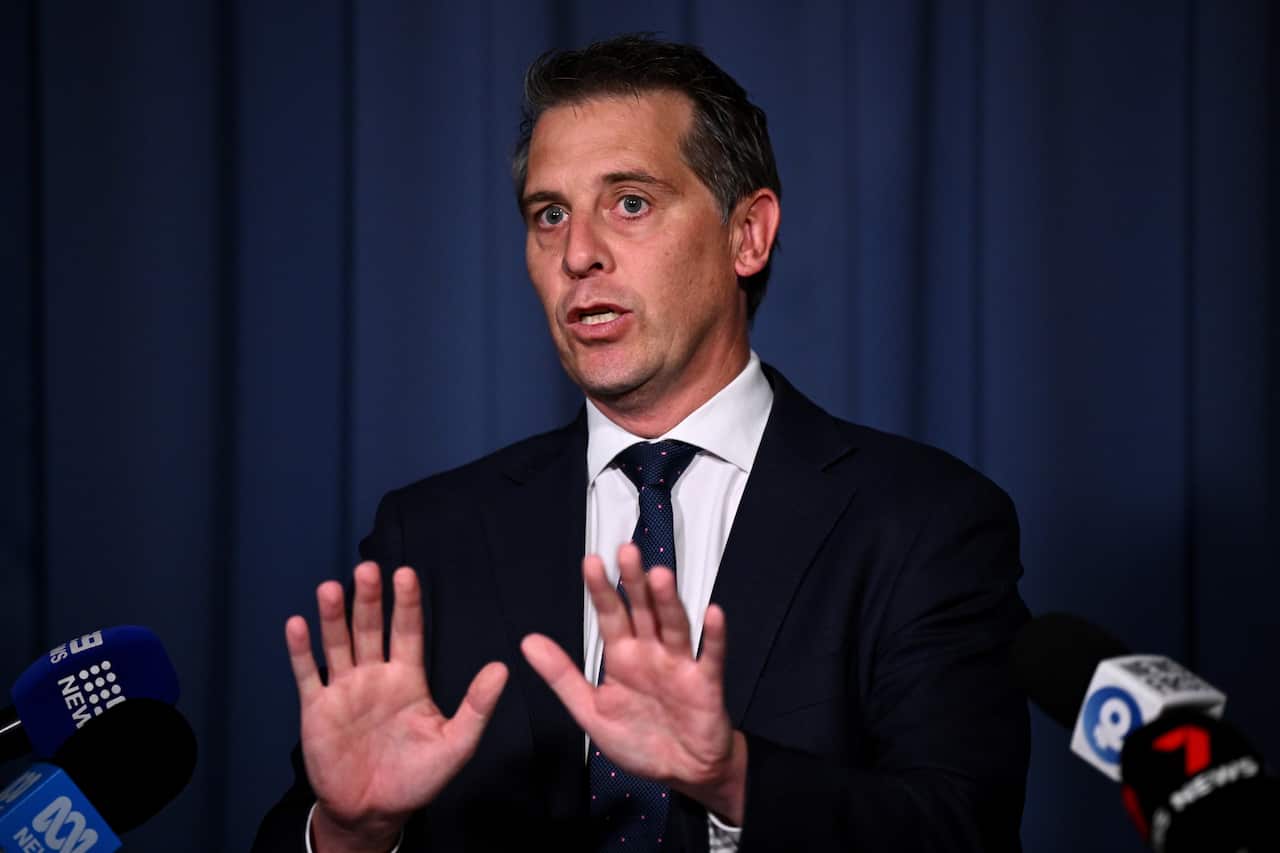 A man in a navy suit and a patterned tie holds his hands up, palms forward, as if to stop something. He stands at a podium with microphones from various news outlets in front of him, speaking with a serious expression on his face.