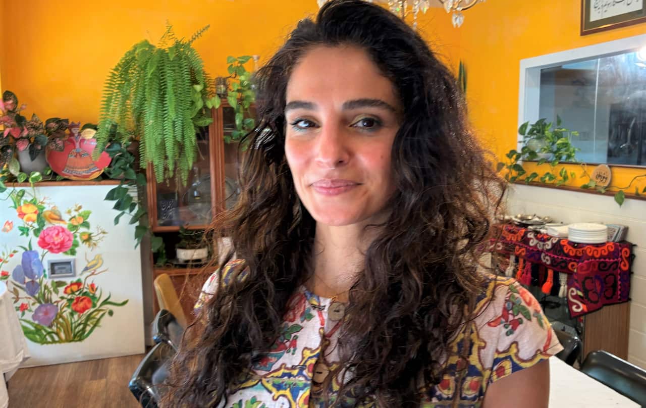 A woman with brown hair wearing a floral dress stands in a restaurant dining room.