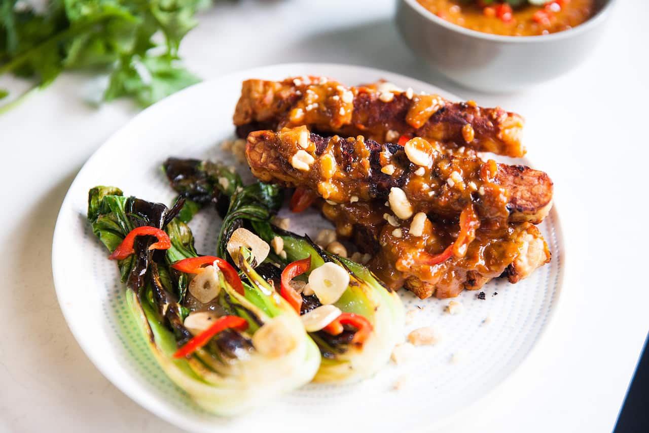 Several strips of crisp tempeh sit on a plate, drizzled with thick brown sauce. Bok choy halves sit alongside. 