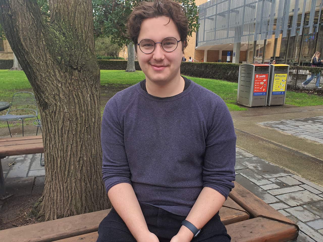 A young man, Atticus Corr, wearing a sweater, sitting by a tree on campus at university.