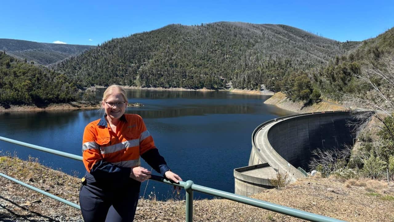a young blonde woman in orange high vis stands by a dam