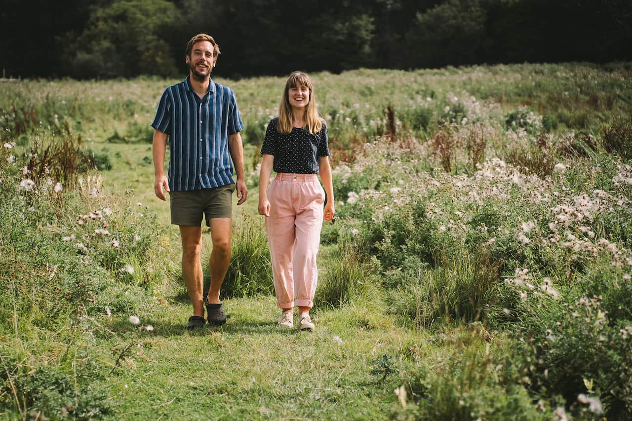 A woman in pink trousers and black shirt walks through a lush green field with a man in a blue striped shirt.