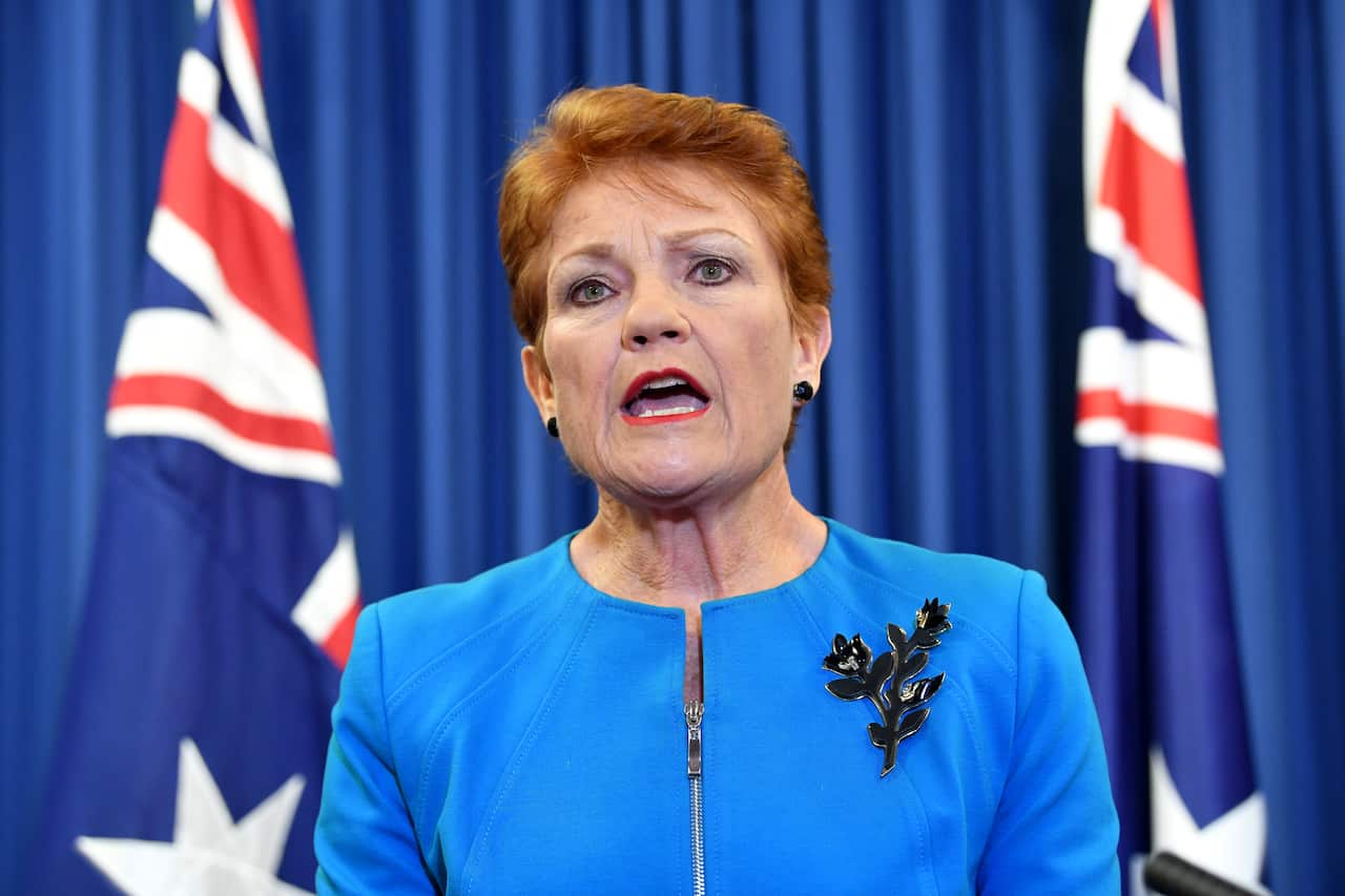 Pauline Hanson speaking in front of a blue backdrop with Australian flags in the background. 