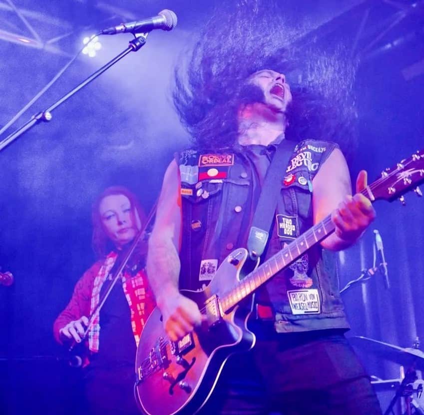 A man in a black denim vest covered in patches plays the electric guitar while flicking his long, dark hair. A woman plays a string instrument behind him