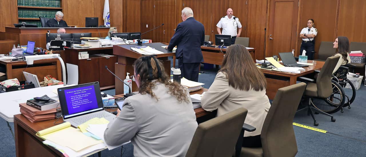 A courtroom during a trial, with wood panelled walls and blue carpet.