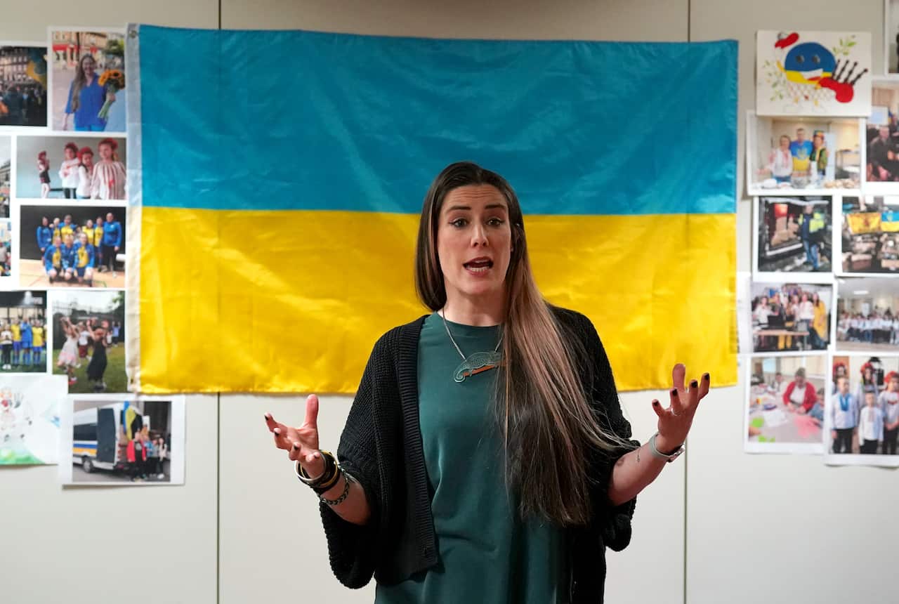 A woman with long hair is pictured mid-speech. Behind her is a Ukrainian flag, and on either side of it, various photos. 