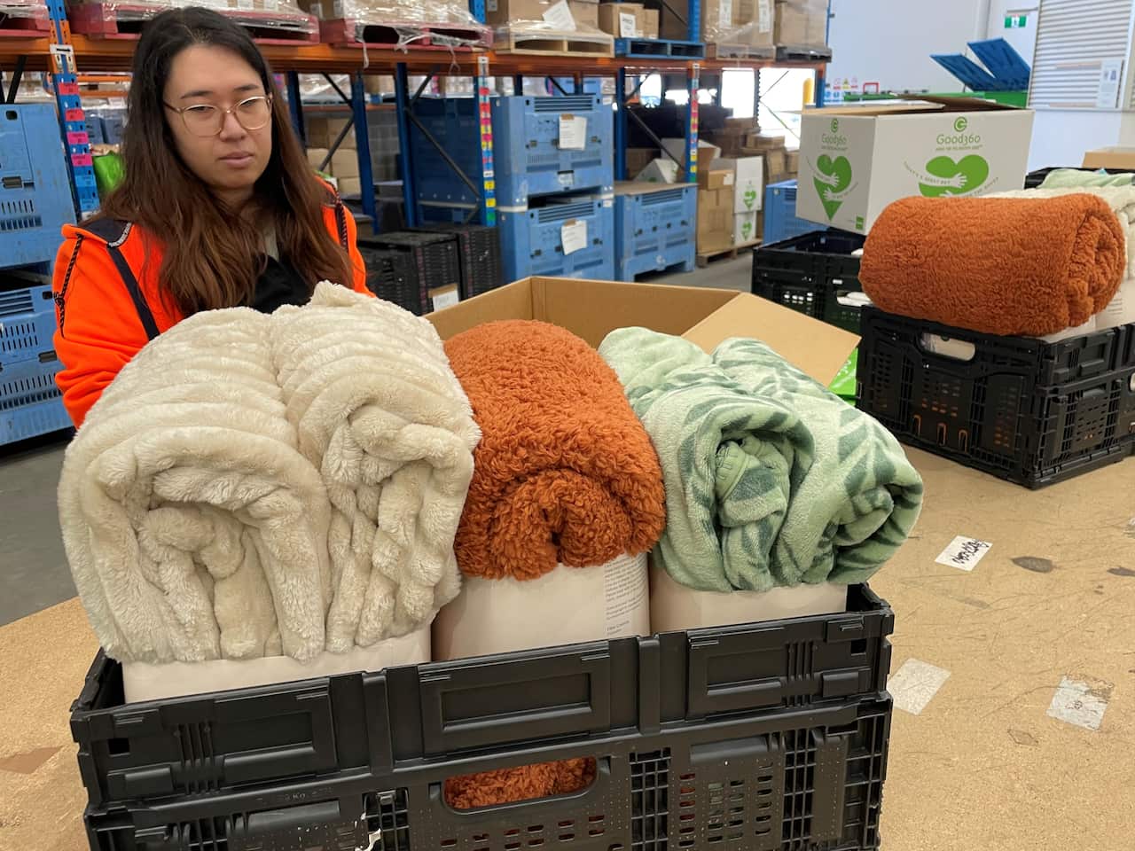 A woman in a warehouse. She is standing behind a black plastic crate containing three quilts.
