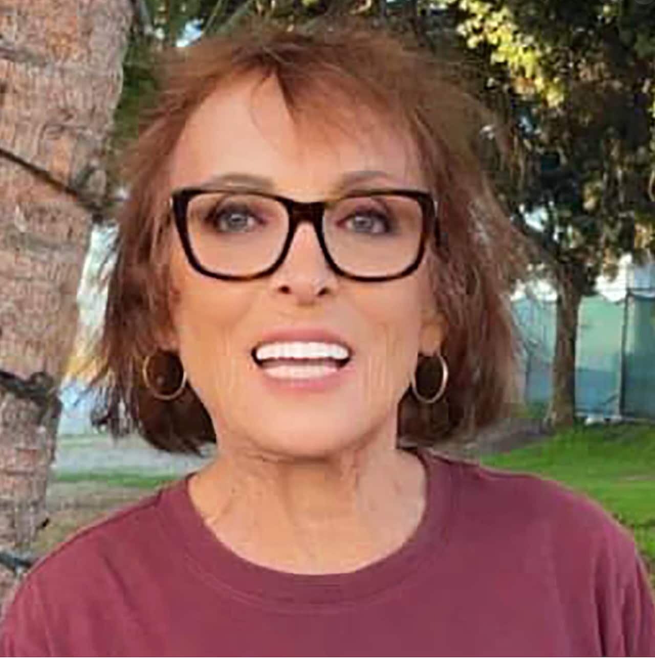 A woman with short hair and glasses, wearing a maroon top, is standing in a garden.