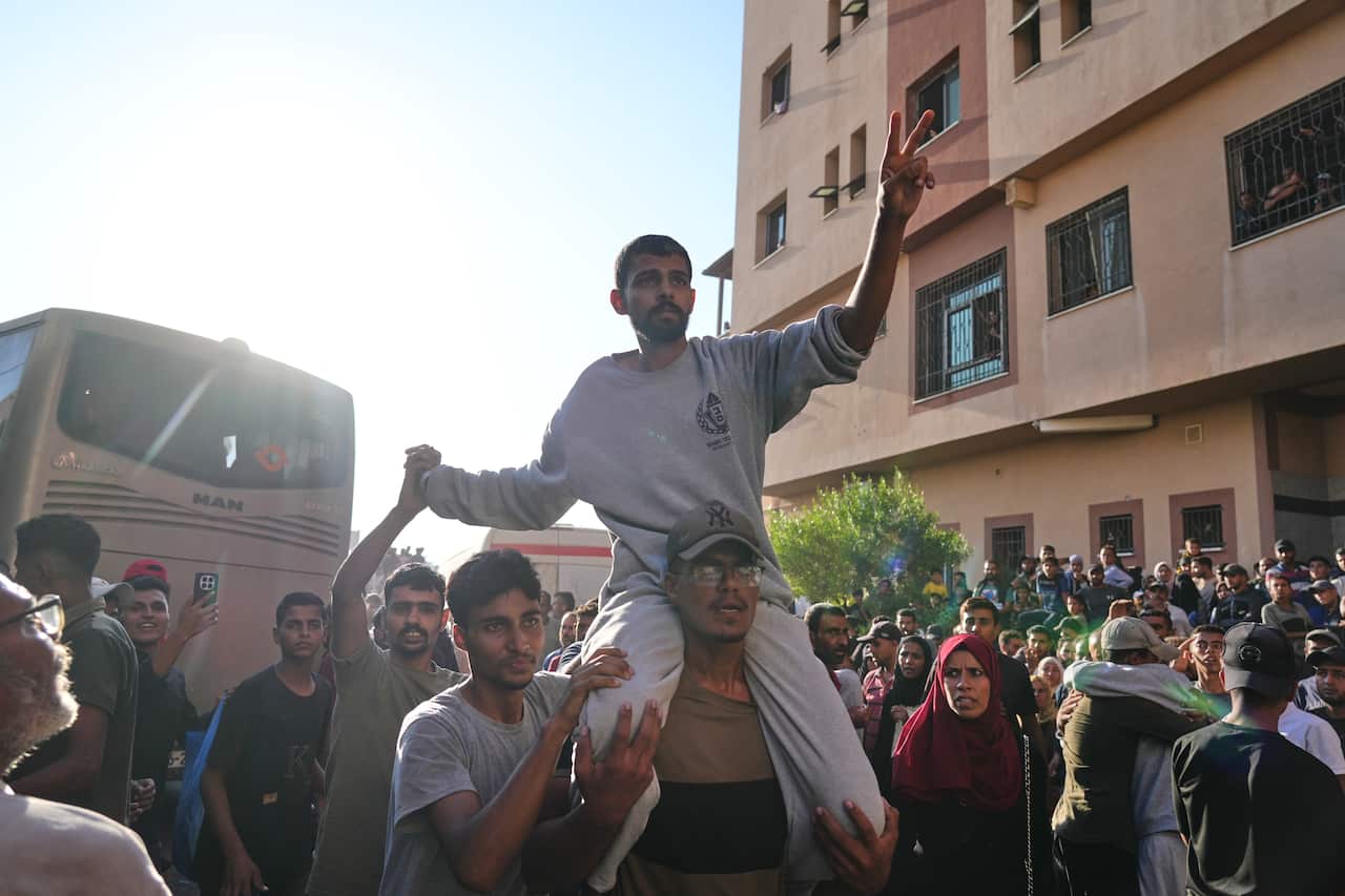 A man flashes a peace sign as he sits on another man's shoulders in a crowd, with a bus in the background.