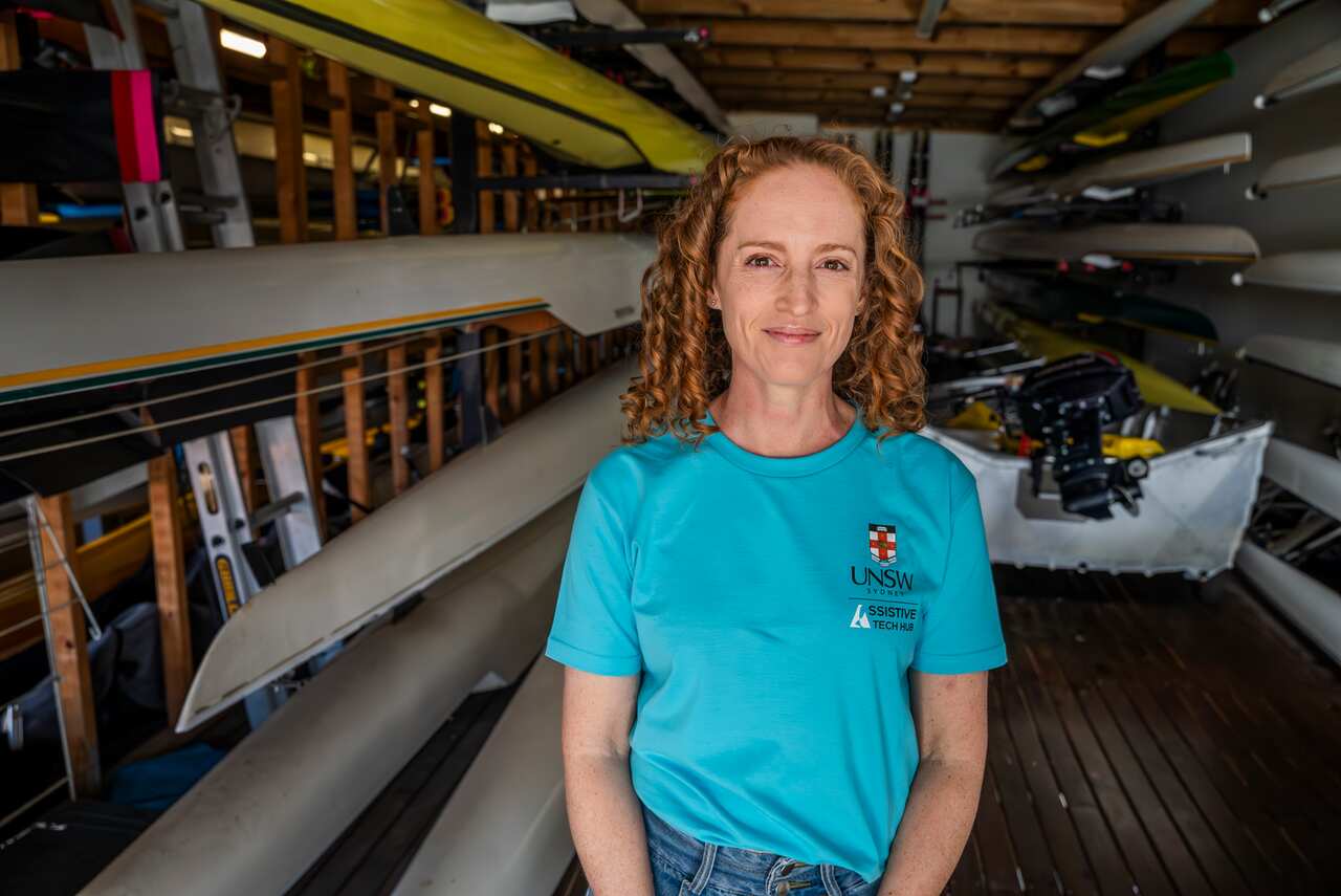 A woman wearing a light blue t-shirt with a university logo stands in a boat shed.