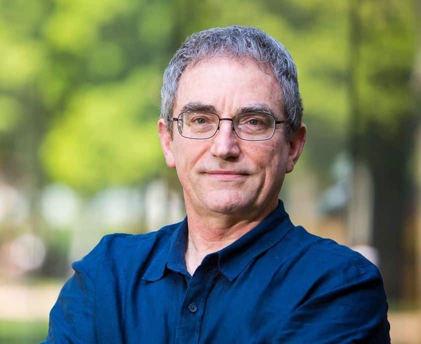 A man with grey hair, glasses and a dark blue dress shirt with out-of-focus trees in the background