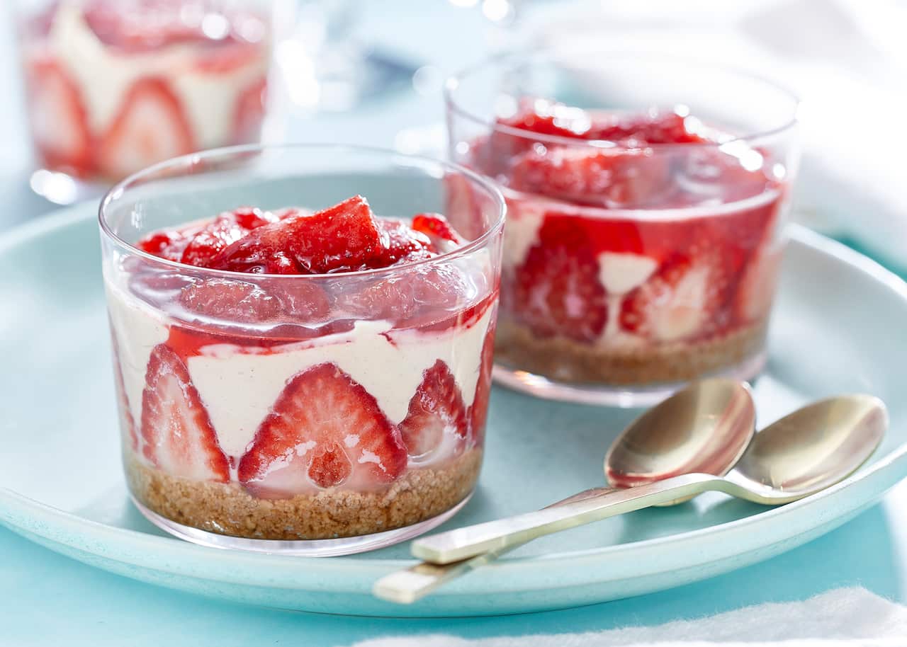 Two dessert serving glasses sit on a wide pale blue plate, along with two spoons. Each glass has a dessert with a crumb base later topped with strawberries and a pale mousse.