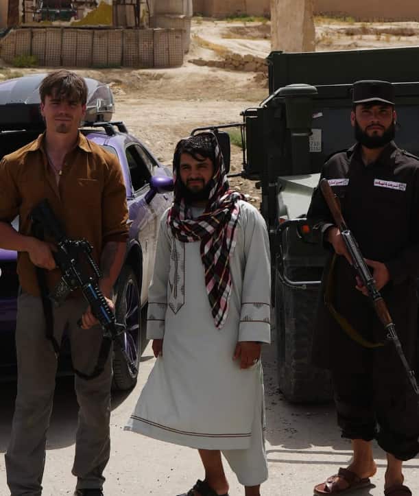 A young white man holding a rifle is standing beside two men: one in an Aghani traditional dress and an armed gunman.