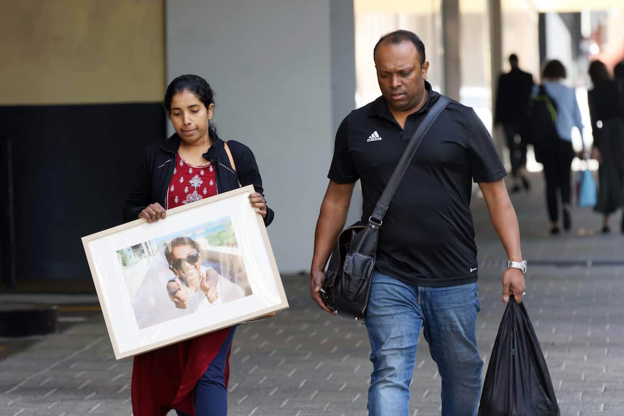A man and a woman walking outside. The woman holds a large framed photograph of a young girl
