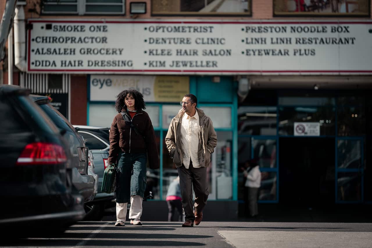 A young woman and a middle-age man walk across a street. A large sign naming multiple businesses including a chiropractor, a noodle restaurant and a hair salon can be seen above the building behind them. 