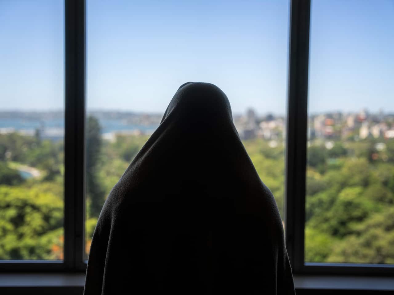 Sarah in a shawl standing at a window looking onto houses and sea.