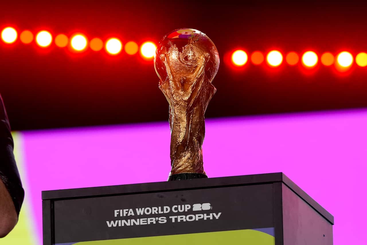 a gold-coloured statuette trophy on a plinth with lights behind it. Writing on the plinth reads FIFA World Cup 26 winner's trophy