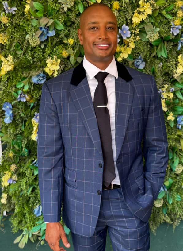 A man in a suit stands in front of a garden wall and smiles.