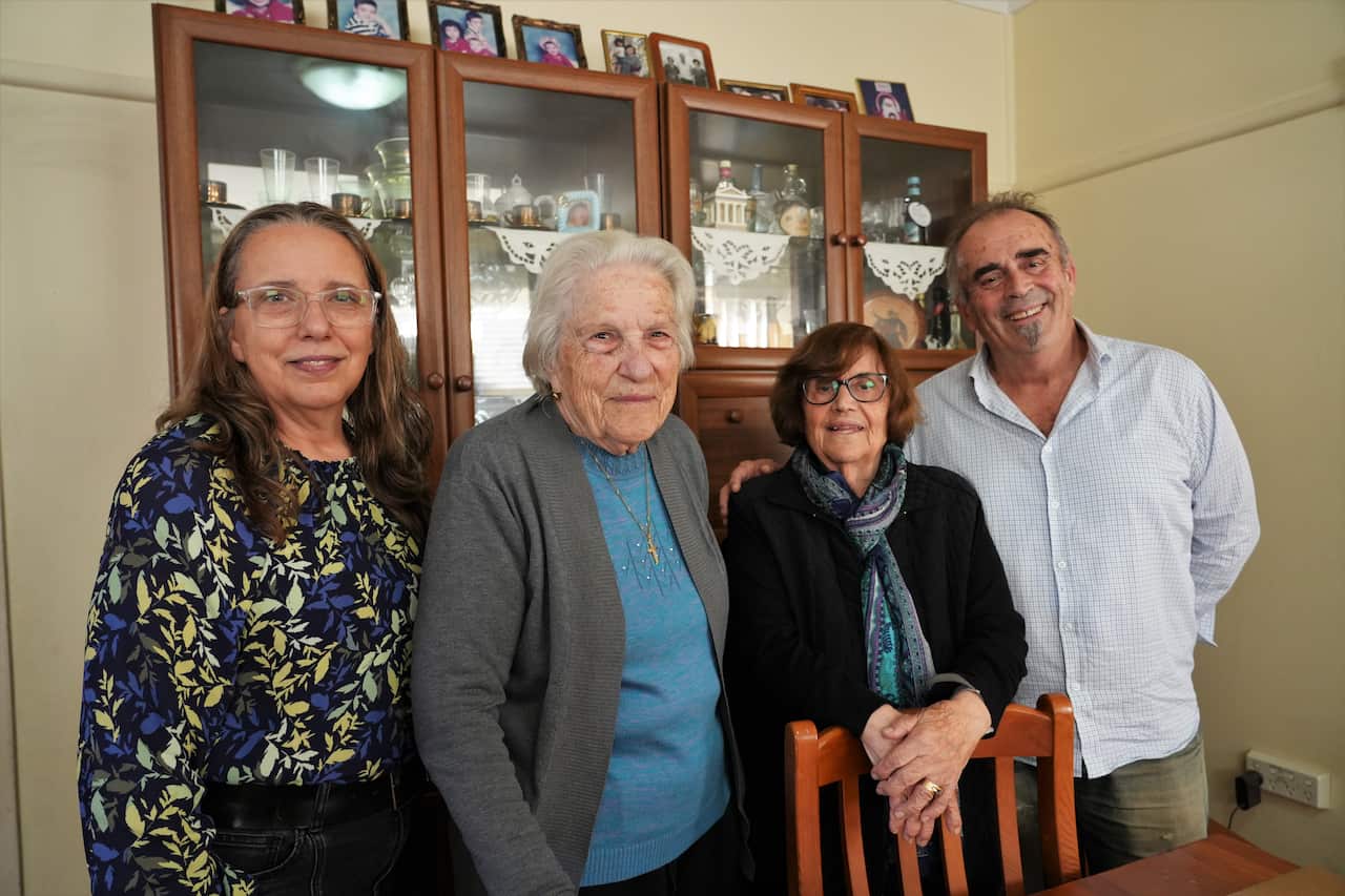 A group of four adults poses indoors in front of a wooden, glass-fronted display cabinet filled with glassware and family photographs. 