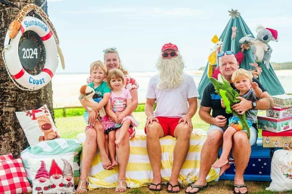 A woman sits with two young children in her lap next to a man dressed as Santa in beach attire, with a man to the right of them sitting with a young boy sitting in his lap holding a toy crocodile 