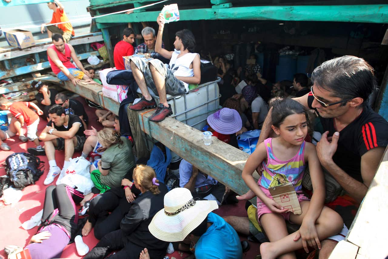 A large, crowded group of passengers, including a man and a young girl in the foreground, are seen sitting tightly together on the decks of a wooden boat.