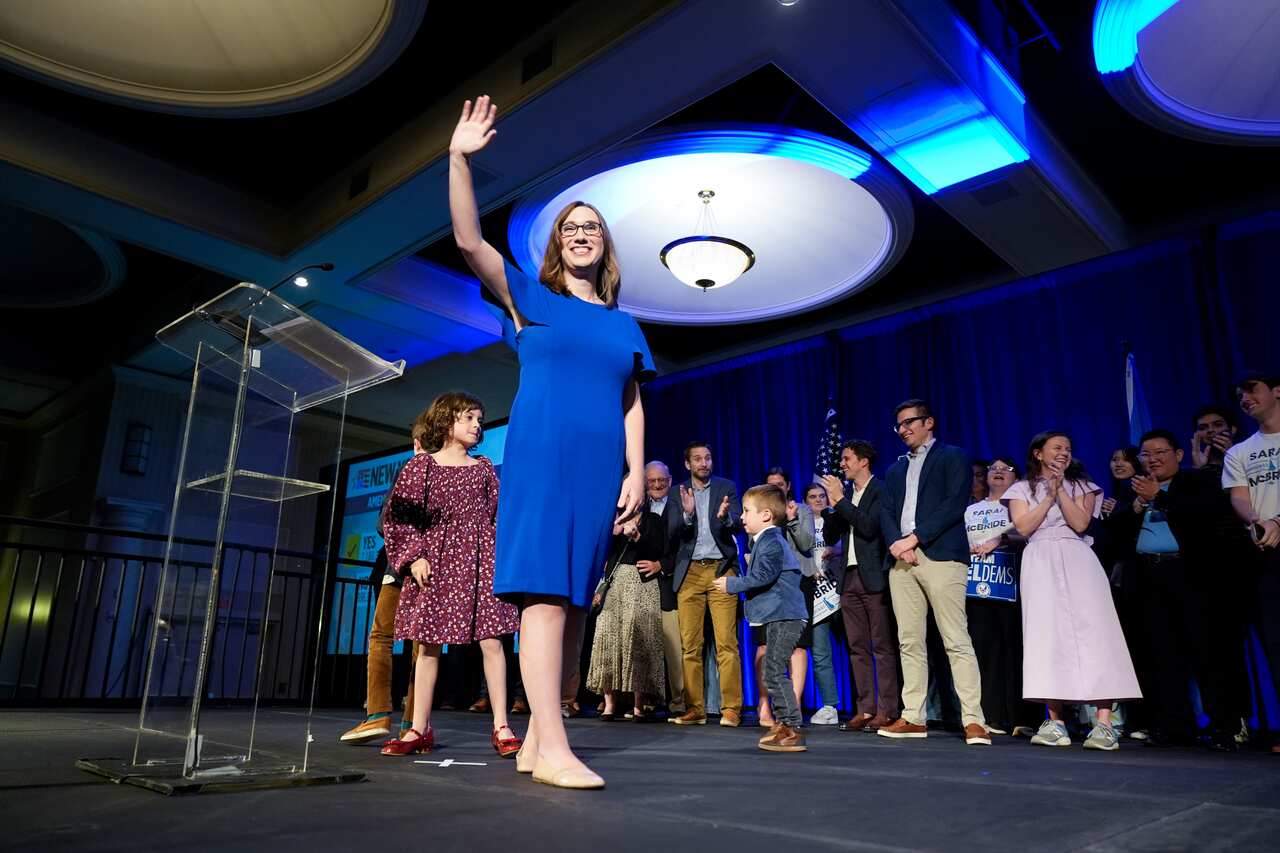 A woman wearing a blue dress waves at a group of people.