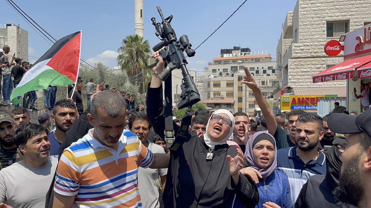 A woman in a black dress and white hijab and wearing reading glasses holding up a rifle in her right hand surrounded by a crowd of people