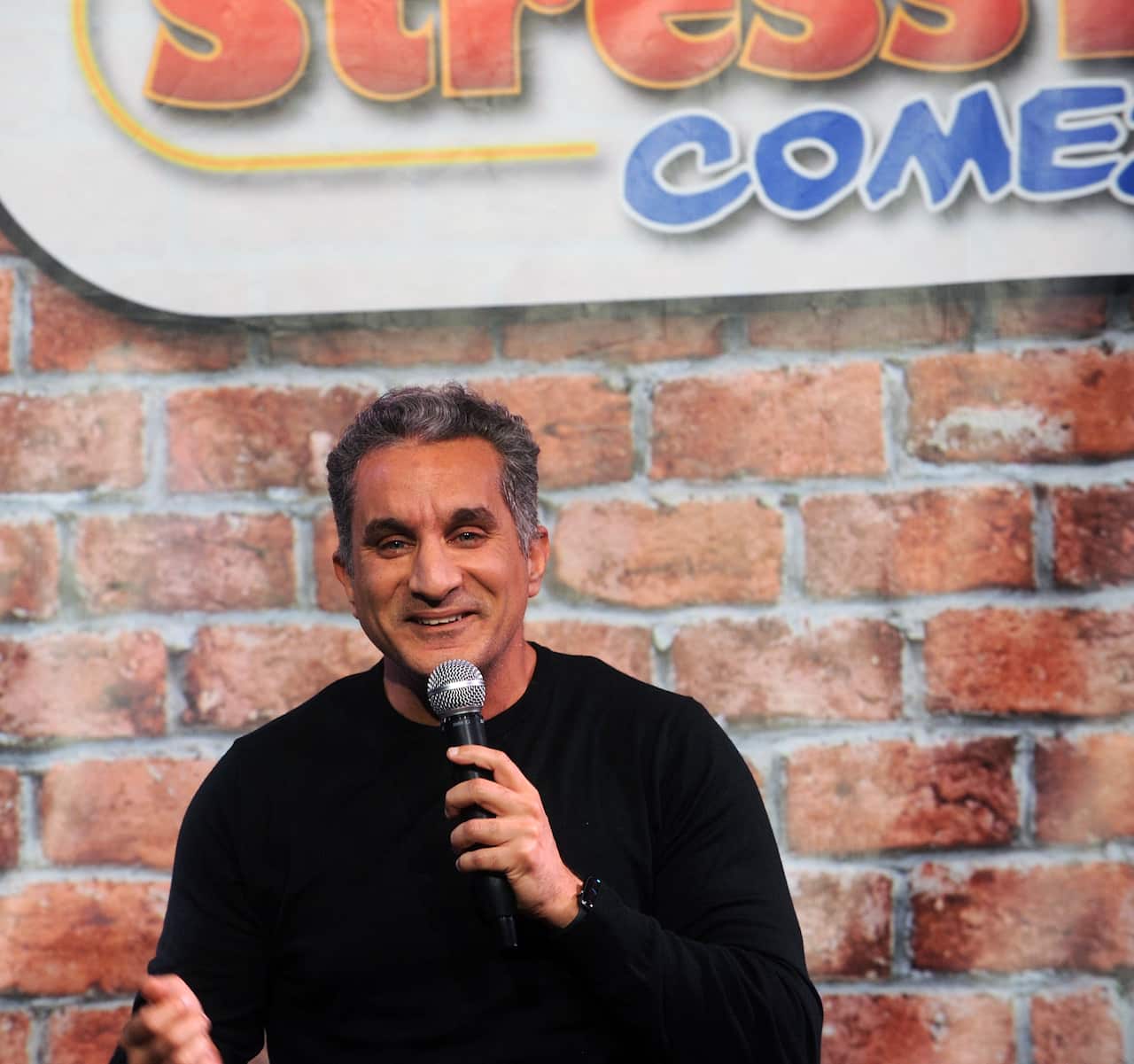 A smiling man in a black jumper holds a microphone while standing against a comedy club backdrop of a brick wall.