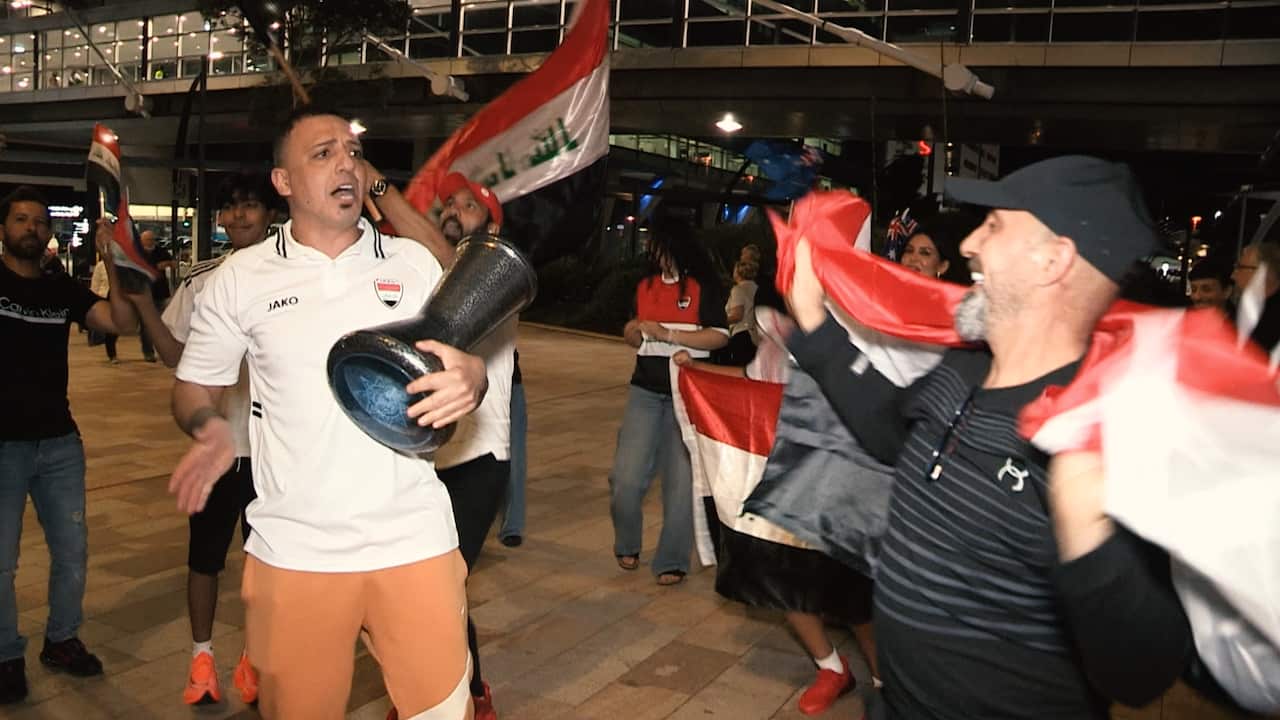 A group of men celebrate at night, smiling and cheering while waving Iraqi flags and playing a traditional drum.