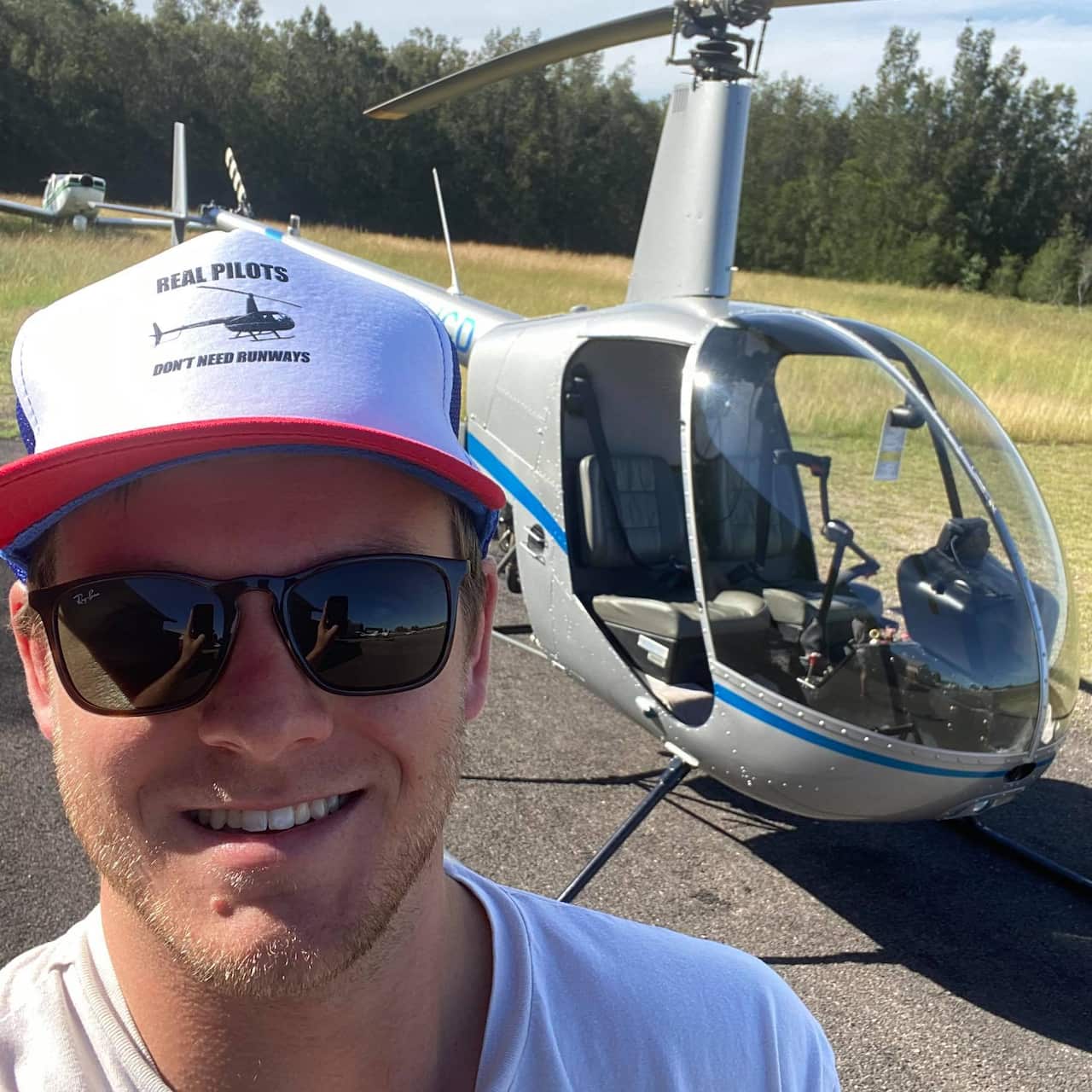 A young man wearing a cap and sunglasses smiling with a helicopter in the background
