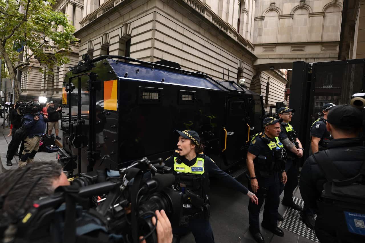 Police officers stand between a prisoner transport vehicle and a crowd of journalists with cameras.