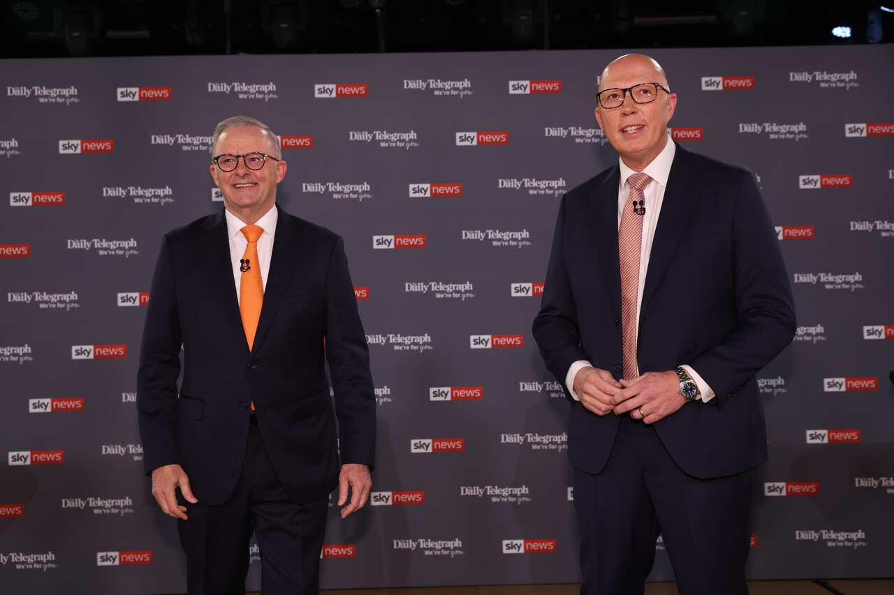 Two men in black suits smile as they stand side-by-side in front of a black background.