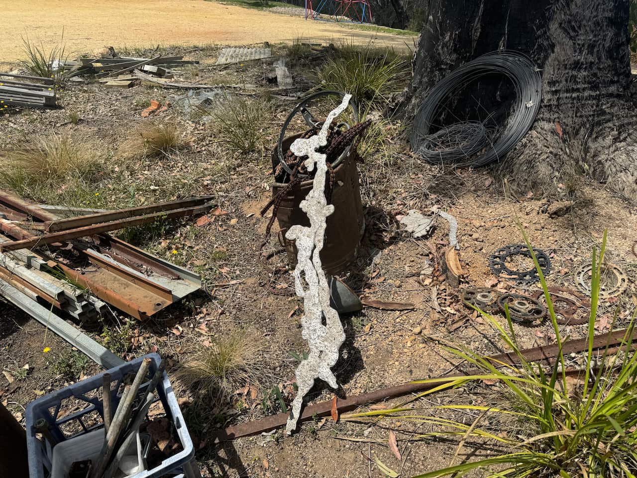 A metal sculpture is lying on the ground next to charred bushes, logs and some other metal parts.