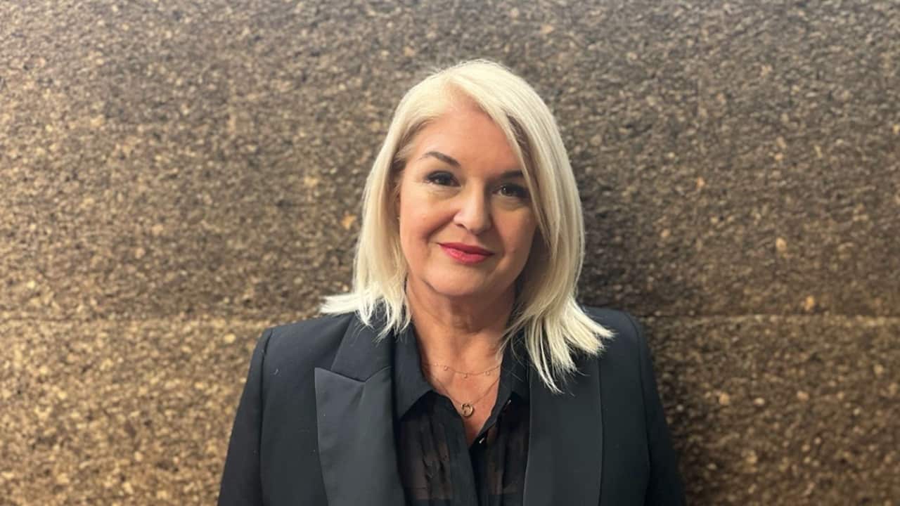 a blonde middle aged woman in a black suit and wearing red lipstick smiles in front of a stone background