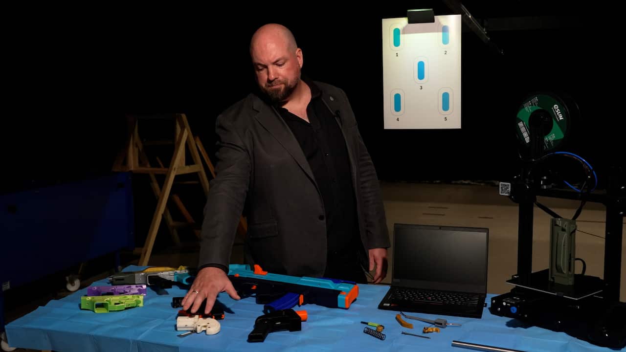 A man in a suit gestures toward 3D-printed guns on a table. 