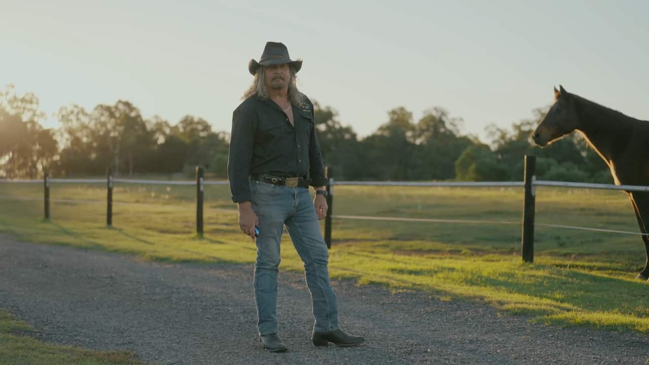 A man in a cowboy hat and boots stands next to a paddock at sunset as a brown horse stares at him from the right.
